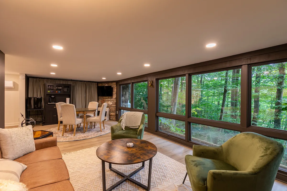 A beautifully lit dining and relaxation area featuring large windows with sweeping views of the forest. The comfortable green armchairs and wooden table highlight a welcoming place for family gatherings, casual meals, or game nights, with forest scenery enhancing the experience.