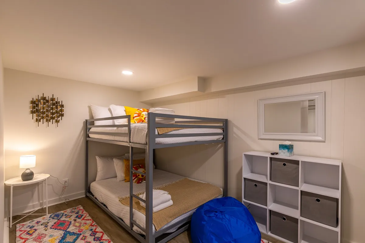 A playful and inviting bunk room with colorful bunk beds, a cheerful rug, a bean bag chair for lounging, and a nightstand with a lamp. The space is designed to accommodate children or groups, providing an atmosphere for both sleep and fun.