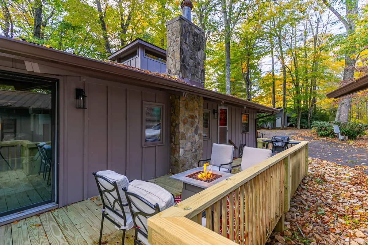 A cozy outdoor area featuring a stone chimney, fire pit, comfortable outdoor seating, and a deck. The seating area is nestled among greenery, providing a perfect setting for socializing, relaxing, or enjoying mountain views with your pet at your side.