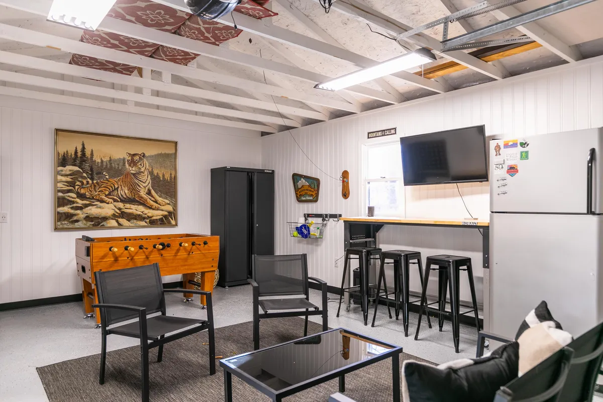 The image highlights a playful garage recreation area with a foosball table, tiger painting, bar stools, a TV, and a refrigerator, creating a perfect setting for game nights and social gatherings. The layout is designed to encourage interaction and fun.