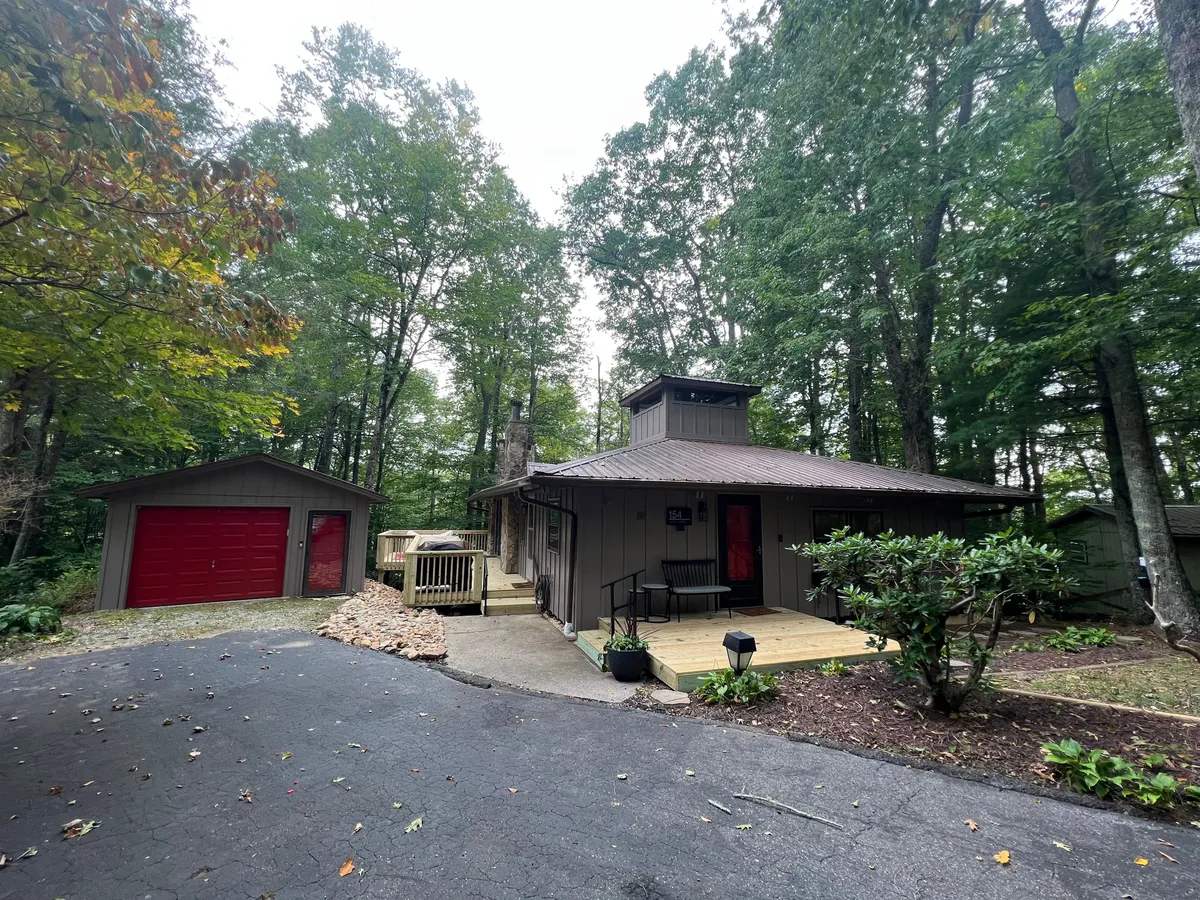 This exterior image showcases the striking red door, unique roof structure, and garage with red accents of Blue Ridge Retreat, surrounded by lush greenery. The wide front porch and natural setting signal an invitation to step outside and begin exploring the property and its natural surroundings.
