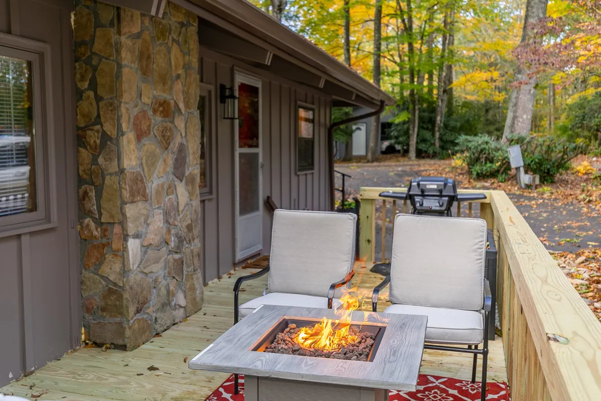 An inviting outdoor scene featuring a fire pit surrounded by two comfortable chairs, a rustic stone wall, and a wooden deck. The area is nestled within a wooded setting, ideal for relaxation and gathering, with warm lighting suggesting the peacefulness of sunset or evening.
