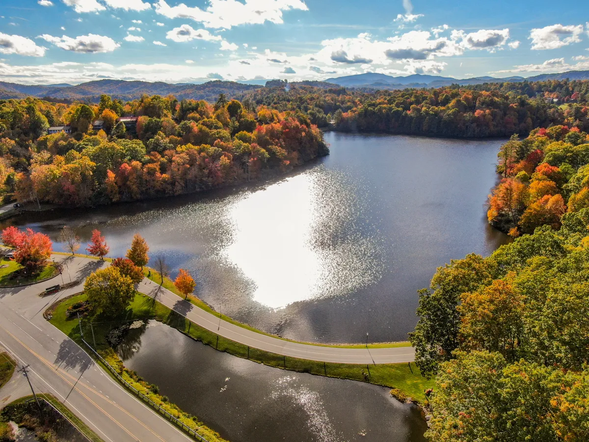 A scenic lakeside view with the shimmering lake at the center, framed by vibrant autumn foliage and a gently curving road. The setting is tranquil, suggesting opportunities for fishing, boating, hiking, and relaxed picnics by the water, while highlighting the beauty of the natural surroundings and year-round enjoyment.
