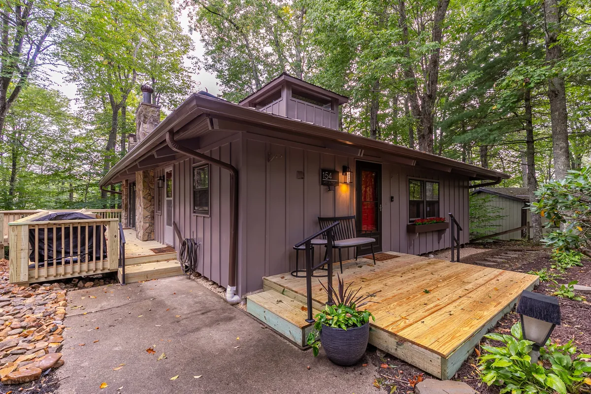 The image showcases the exterior of a modern cabin surrounded by lush green foliage, featuring a spacious wooden deck, a striking stone chimney, and a welcoming red door. The scene is set during sunset, casting a warm, inviting glow over the porch—perfect for gatherings or quiet moments with morning coffee amid nature.