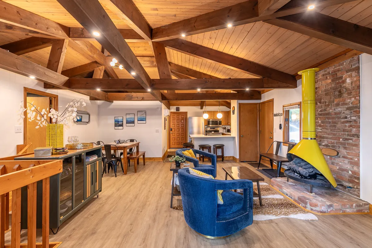 This image showcases a bright, open-plan living and dining space with exposed wooden beams, a vivid yellow chimney stove, a comfortable seating area, and a welcoming dining table. The large windows allow abundant natural light to fill the room, highlighting both the modern finishes and warm, communal layout.
