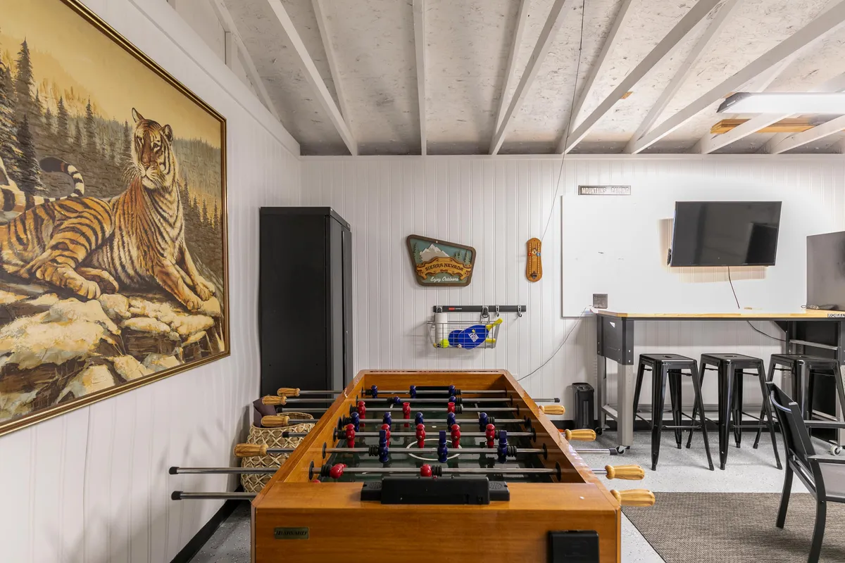 This image displays a lively recreation room featuring a foosball table at the center, a bold tiger painting on the wall, a sleek bar area with stools, and a TV mounted nearby. The setup is purposely casual and geared toward social interaction and entertainment.
