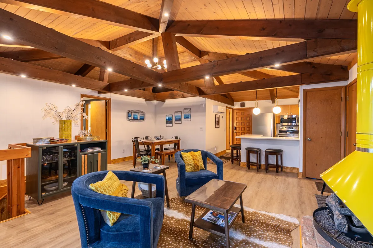 This view features open living and dining areas with exposed wooden beams, a prominent yellow fireplace, stylish blue armchairs, and a spacious dining table. It radiates warmth and invitation, with plenty of space for group gatherings or intimate conversations.