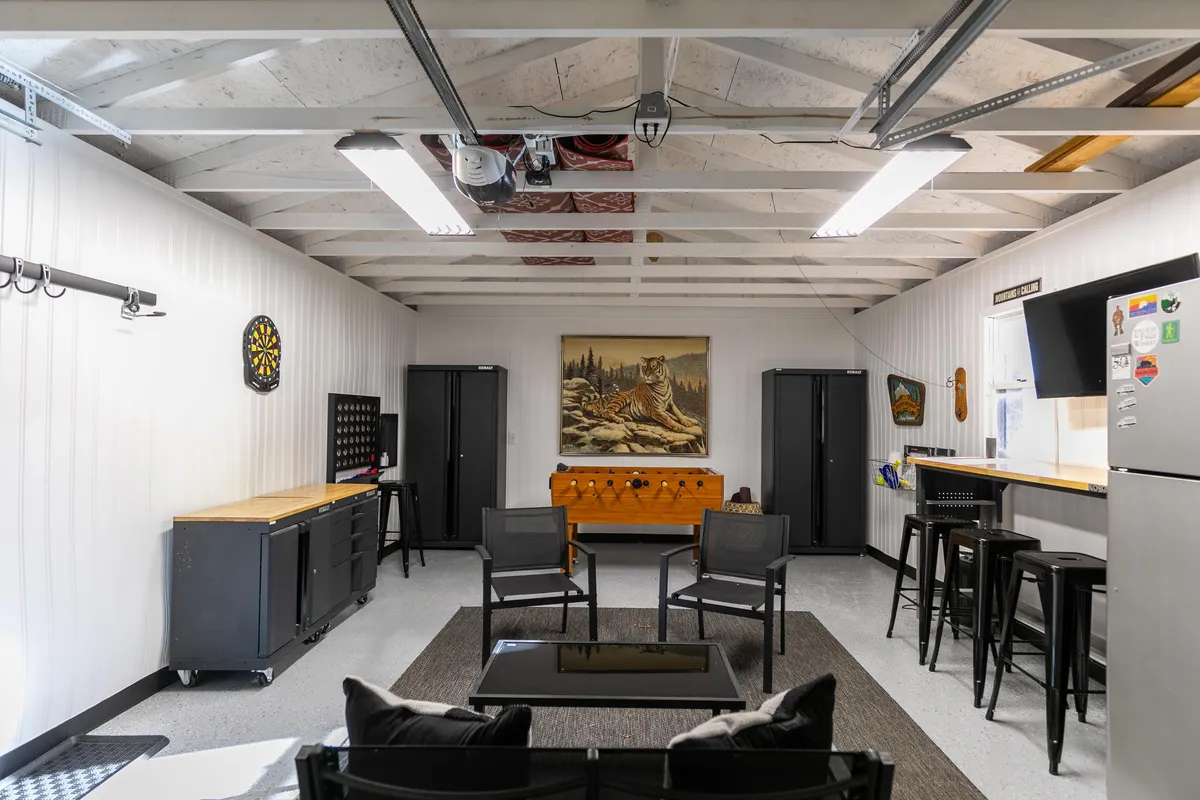 This image focuses on the garage's dartboard area, complemented by a wooden chest, tiger artwork, bar stools, and a cozy seating zone. The space is arranged to facilitate both friendly games and relaxed socializing.