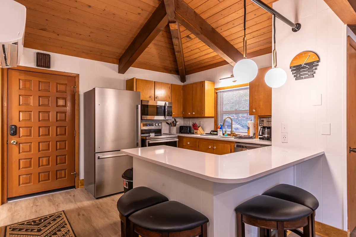 A modern kitchen scene with a central island countertop, wooden ceiling beams, stainless steel appliances, and a bright window offering ample light. The space feels welcoming and ready for group cooking or casual meals.