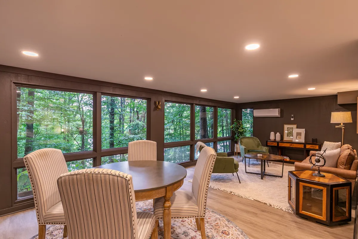 This image showcases a spacious dining table set near large windows that overlook lush greenery outside. The comfortable seating area and abundance of natural light create an inviting atmosphere for sharing meals, morning coffee, or casual conversations with a scenic forest backdrop.