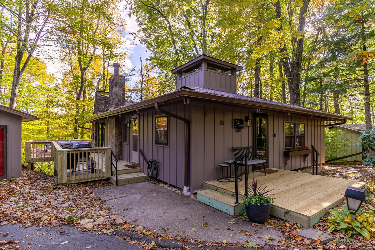 A welcoming front porch set against tall trees, with a visible stone chimney and a wooden deck. The space is surrounded by nature, evoking tranquility and offering a seamless transition from indoors to the outdoors, perfect for relaxing or preparing for an adventure with your pup.