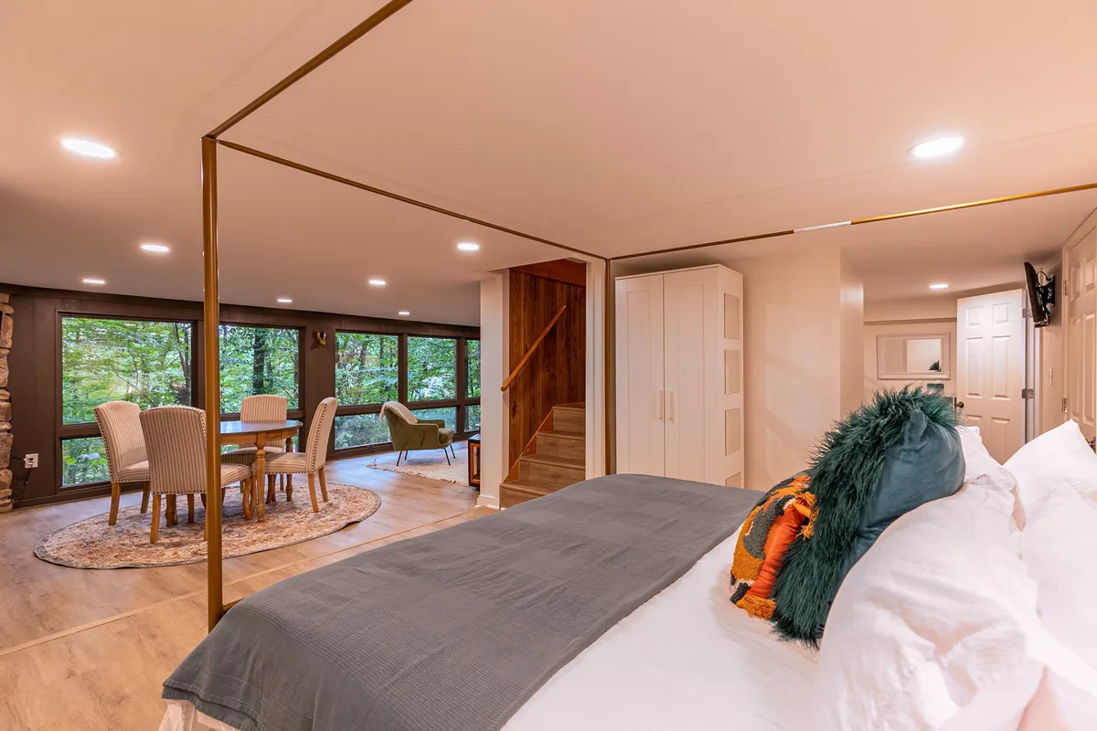 A cozy combined space with a bed featuring decorative pillows, a dining table set for four, and expansive windows that showcase lush greenery outside. The atmosphere is intimate and inviting, perfect for relaxing, enjoying meals, or taking in calming nature views.