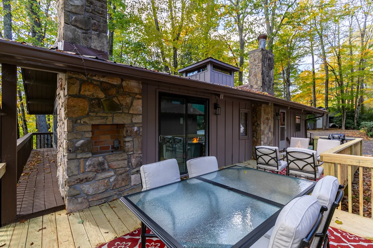 An exterior view of a wooden deck featuring a glass-top dining table, surrounded by trees and bordered by a stone chimney. The deck provides a seamless transition between indoor comfort and outdoor adventure, ideal for dining, relaxing, and socializing after a day of exploration.